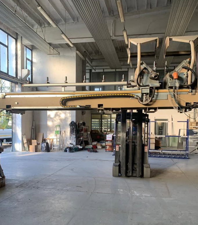 Un lungo braccio industriale beige, montato su una base per carrello elevatore, si trova all'interno di un grande capannone-officina aperto.