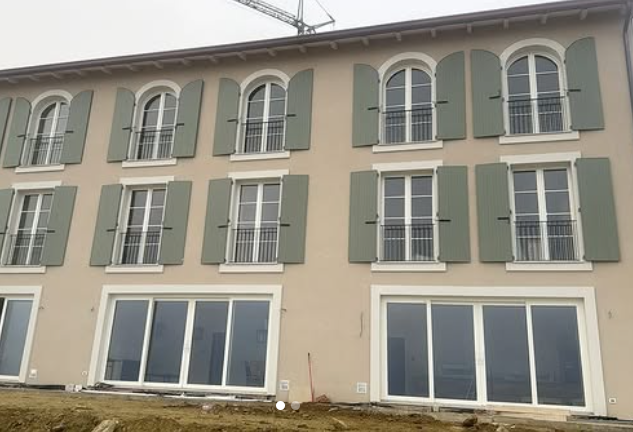 Un edificio di tre piani di colore chiaro con finestre ad arco, persiane verde salvia e grandi porte scorrevoli in vetro al piano terra.