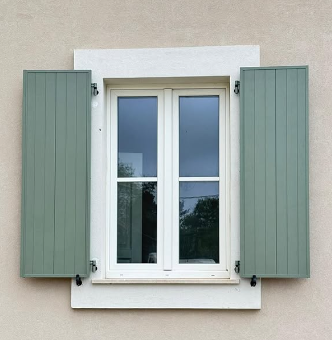 Una finestra a doppio vetro con infissi color crema e persiane verticali verde chiaro, incastonata in una semplice parete beige.
