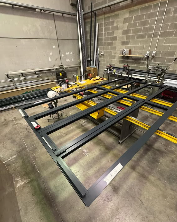 In una luminosa officina industriale, un telaio di una finestra o di una porta in metallo nero è in fase di costruzione su un banco da lavoro.