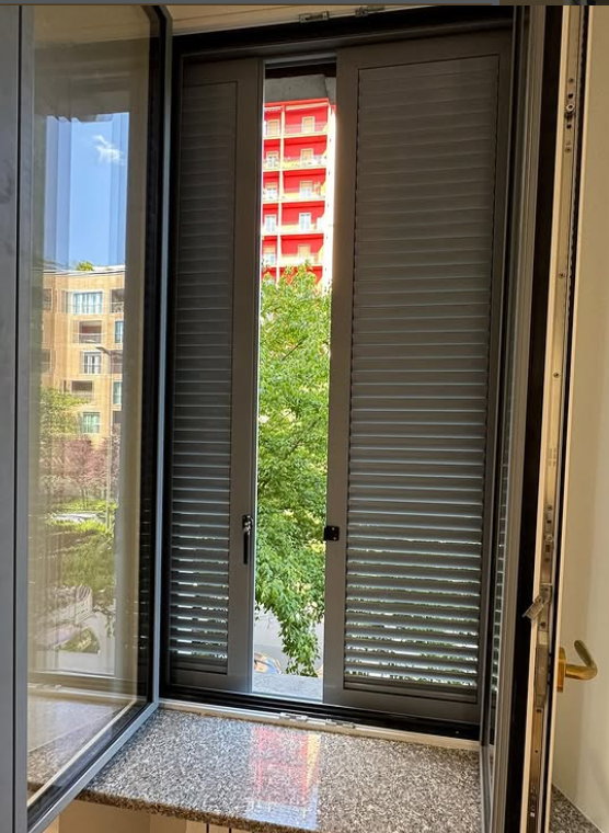 Una finestra aperta con persiane a lamelle grigio scuro, che si affaccia su un albero e un edificio rosso.