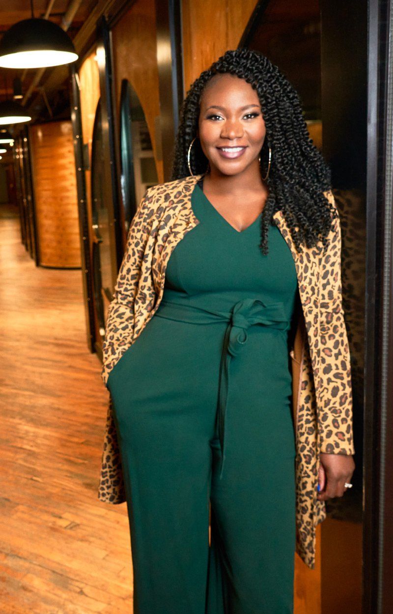 Woman in green jumpsuit and animal print jacket smiles in a hallway.