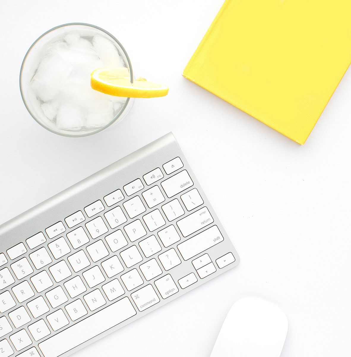 Keyboard, glass of water with lemon, yellow book, white mouse on white surface.