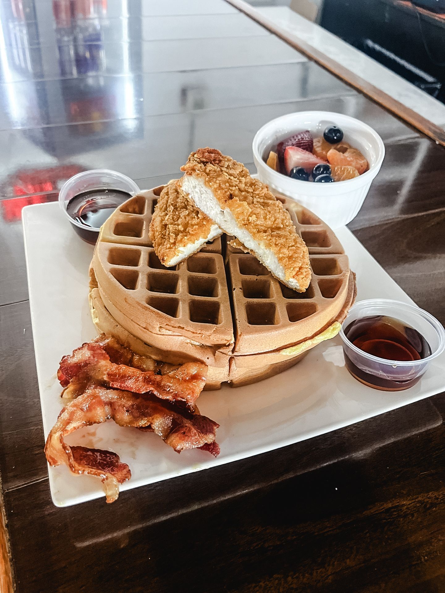 A plate of food with waffles , bacon , syrup and fruit on a table.