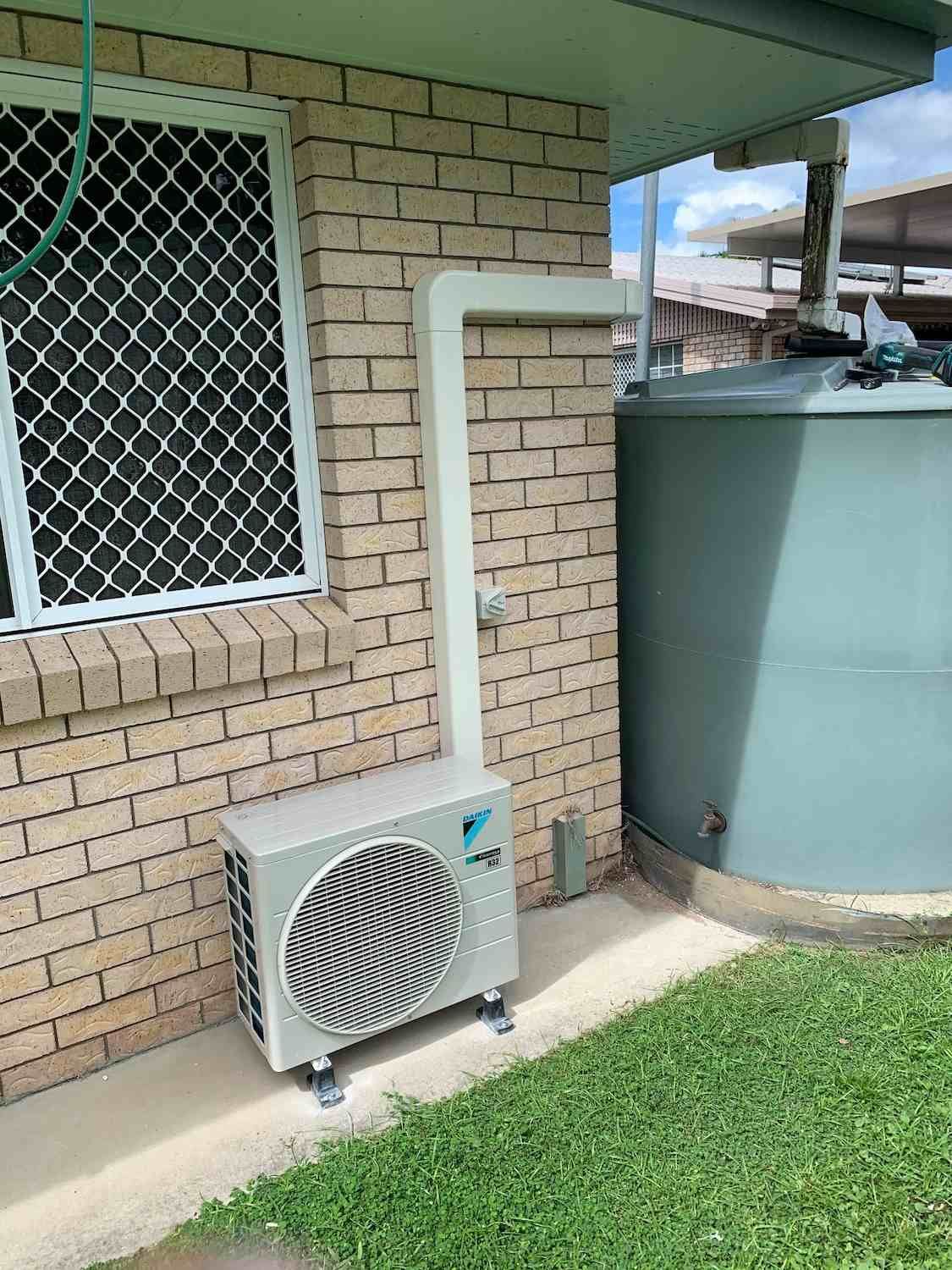 A White Air Conditioner Is Sitting on The Side of A Brick Building Next to A Water Tank — McGuire Air & Refrigeration In Walkerston, QLD