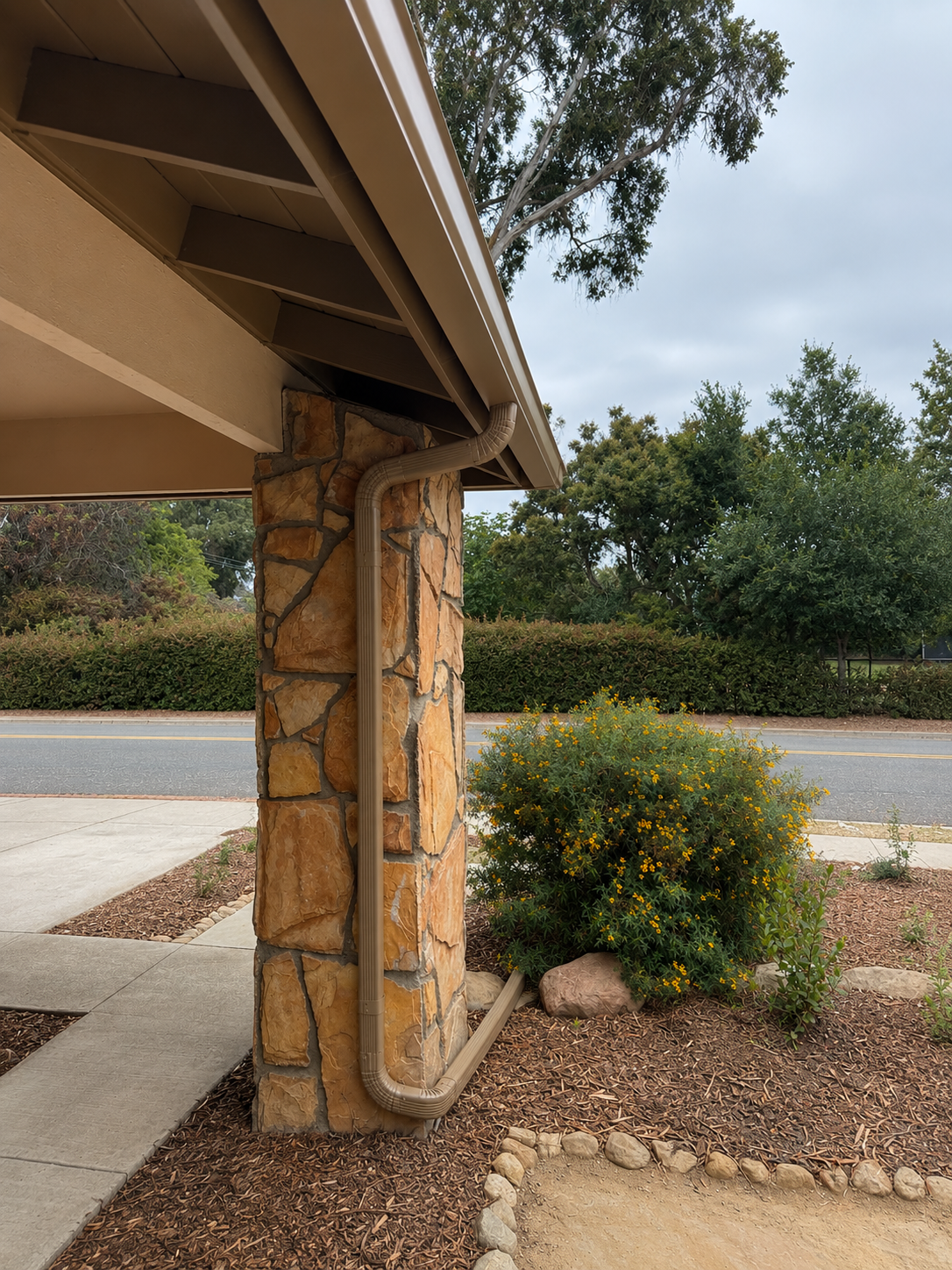Covered walkway with stone pillar beside a sidewalk and landscaped shrubs near a street.
