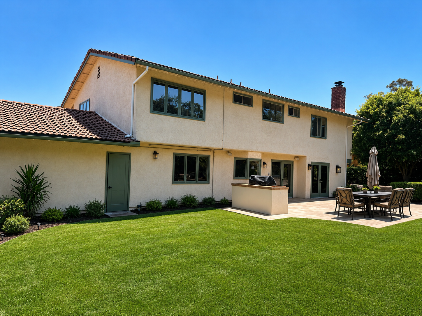 Two-story beige house with a green lawn, patio table, and umbrella under a clear blue sky