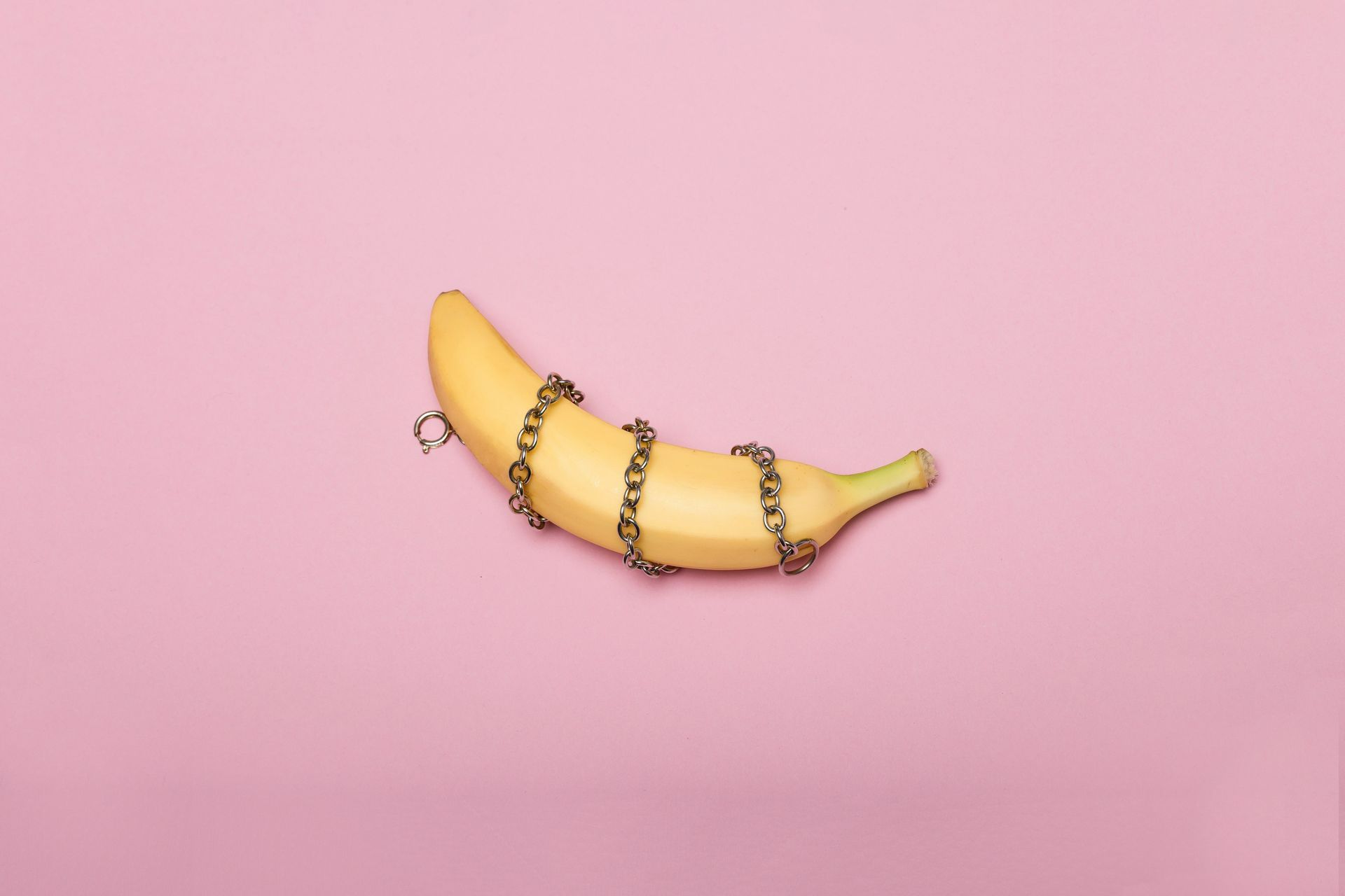 A banana is wrapped in chains on a pink background to symbolize BDSM.