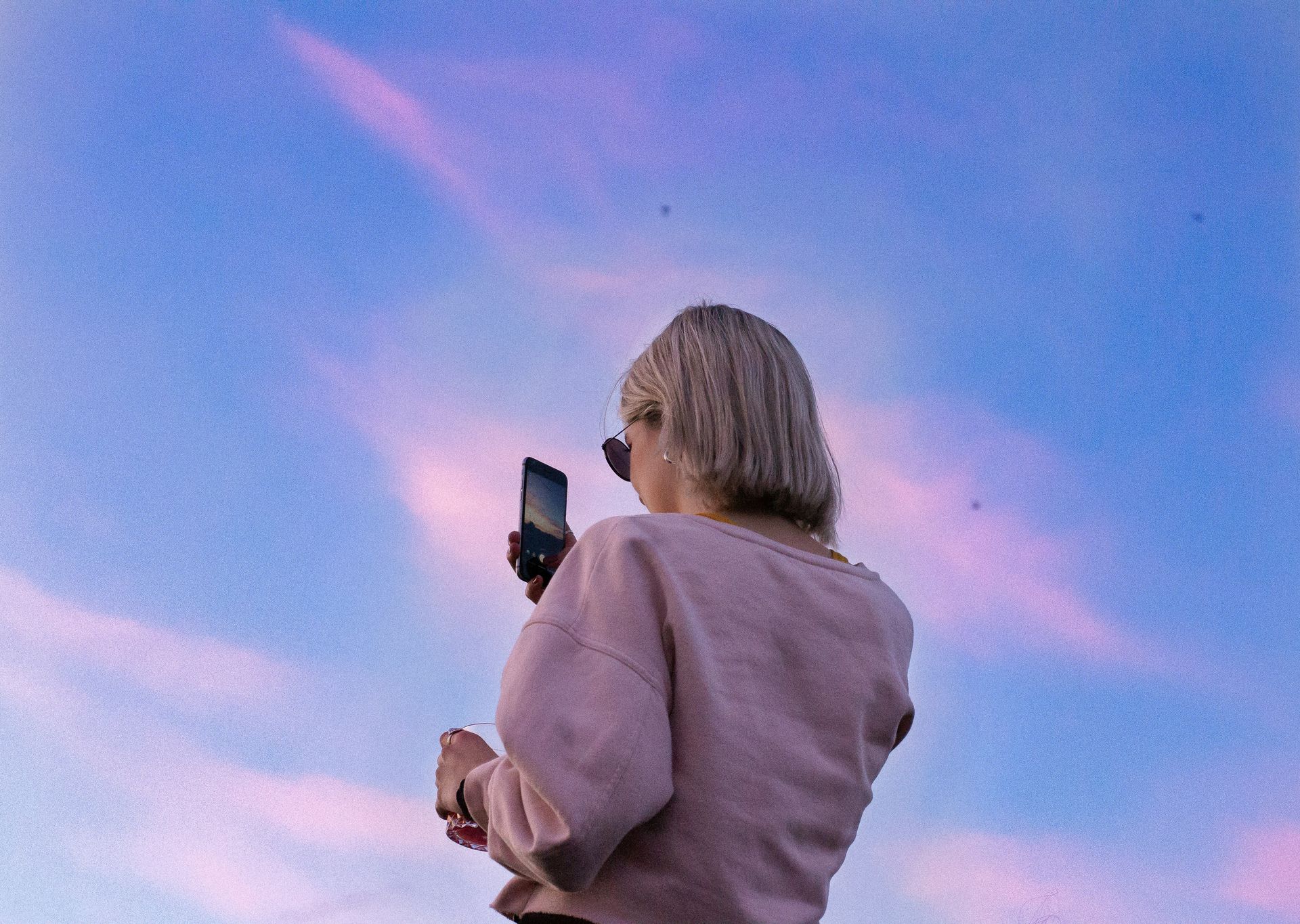 A woman in a pink sweatshirt scrolls on her phone in front of a pink and blue sky. 