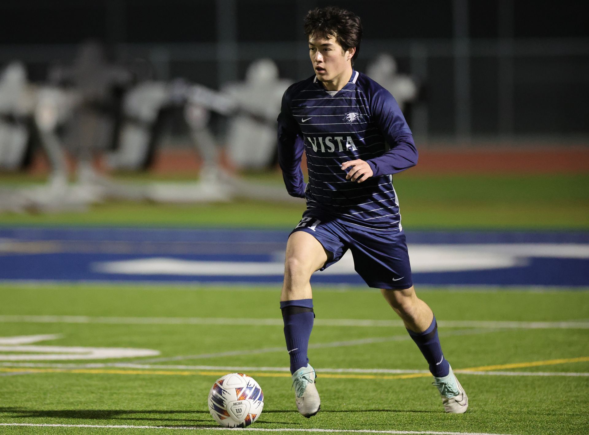 Soccer player in blue uniform dribbling a ball on a green field at night, stadium lights on. Vista Athletic Boosters