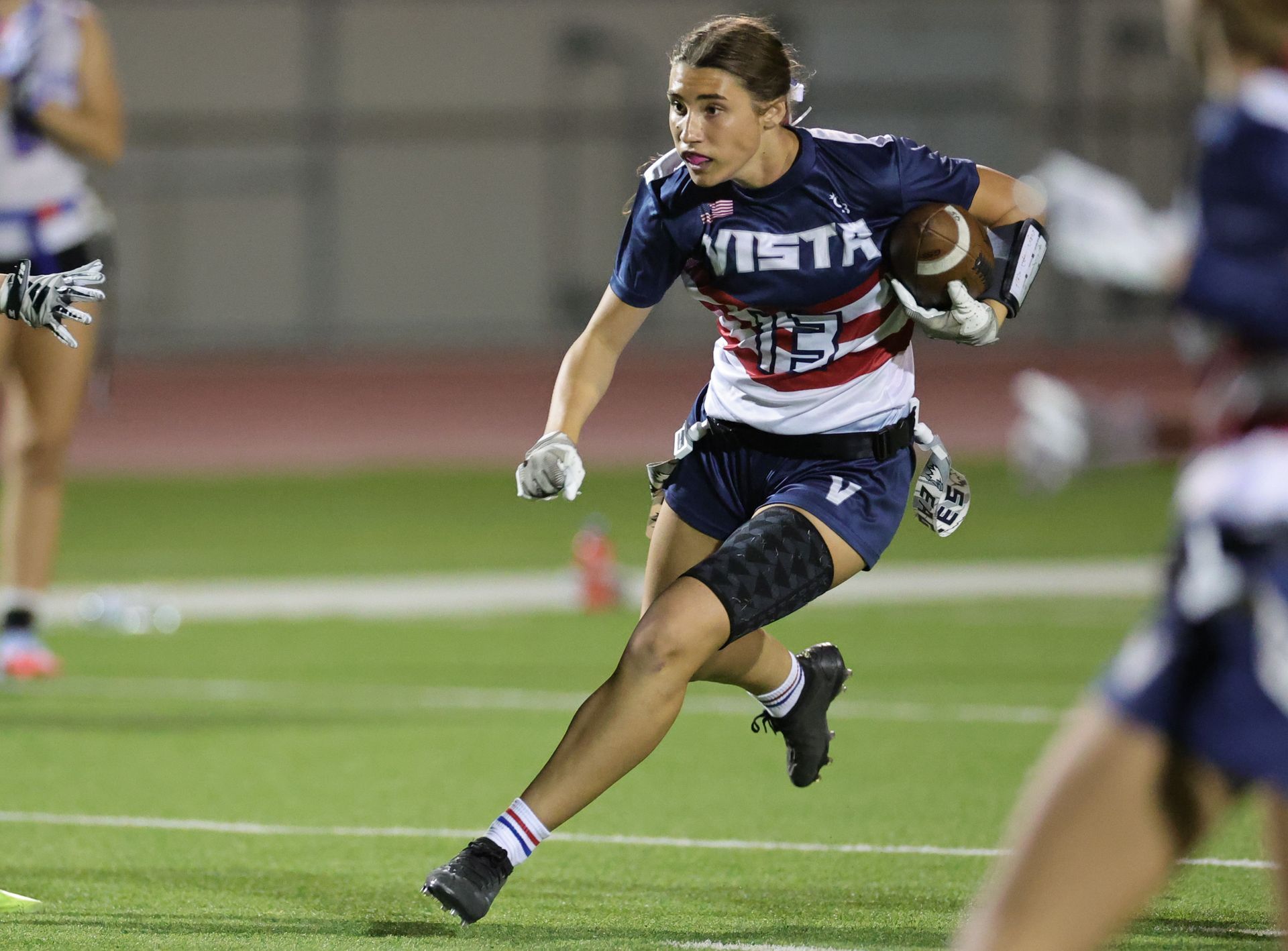 Female flag football player running with ball on a green field. Wearing blue and white uniform. Vista Athletic Boosters