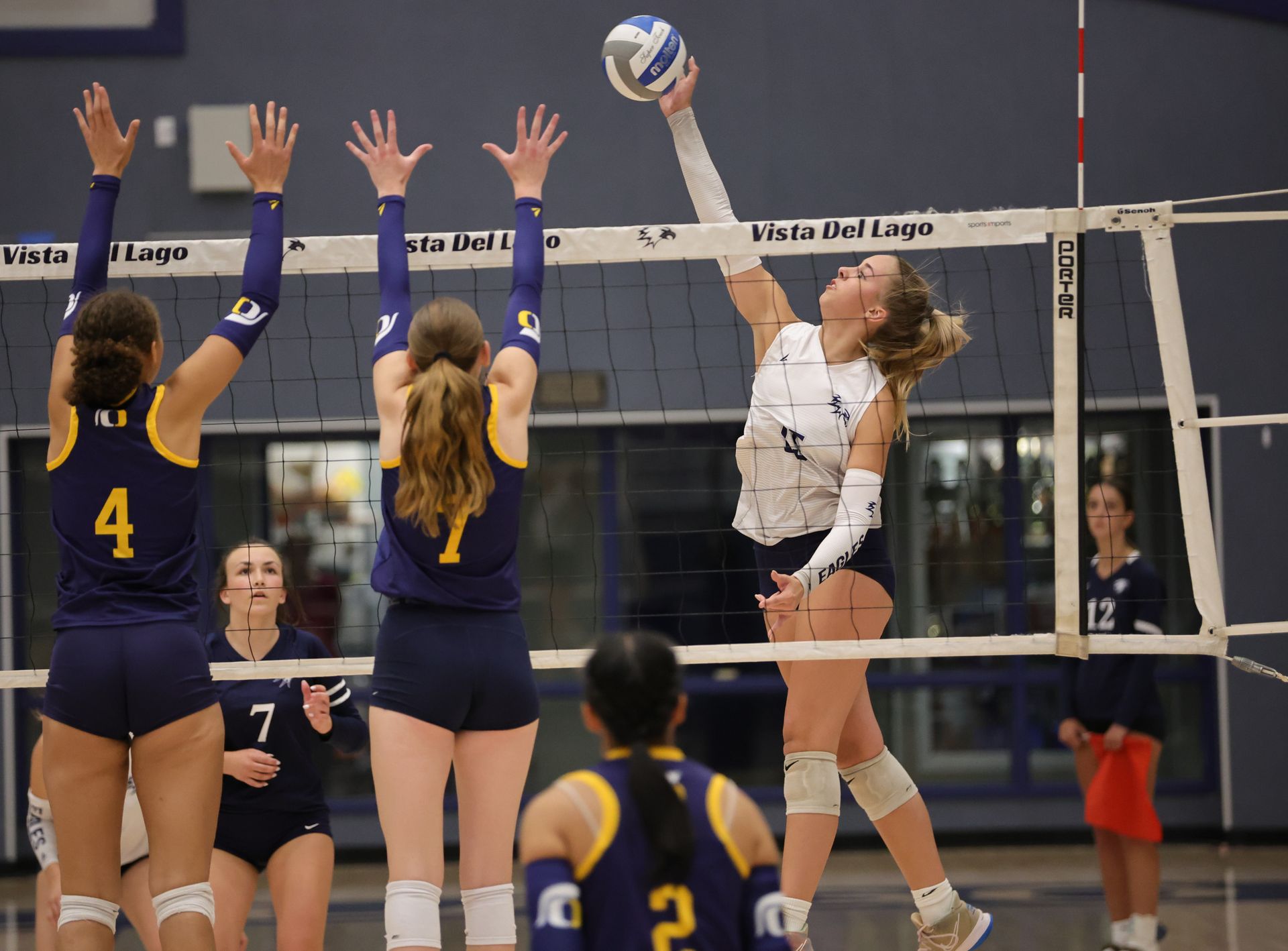 Volleyball player spikes the ball over the net during a game, two players block Vista Athletic Boosters
