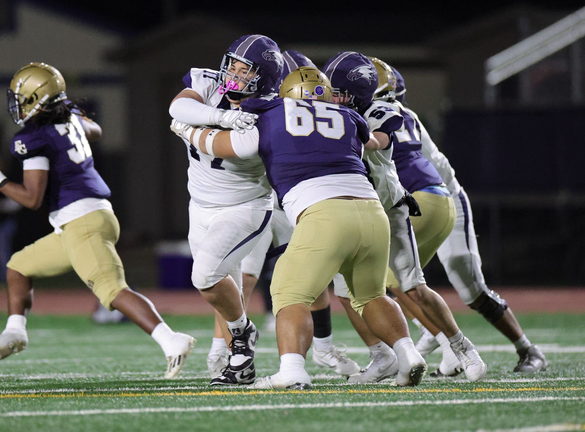 Football players in white and gold uniforms, tackling player in purple and white uniform on green field.