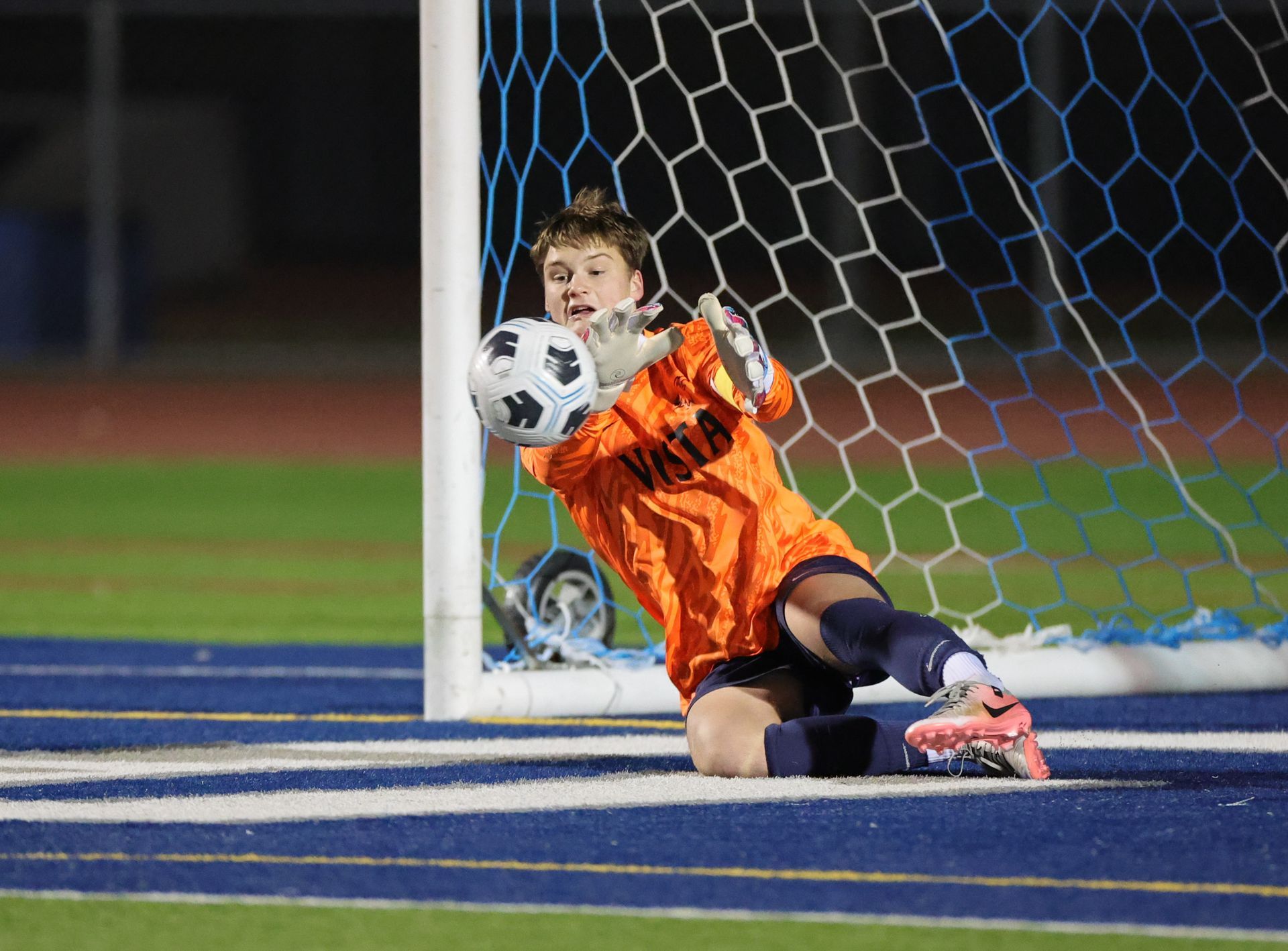 Soccer goalie in an orange jersey dives to catch a soccer ball near the net, on a blue and green field at night. Vista Athletic Boosters