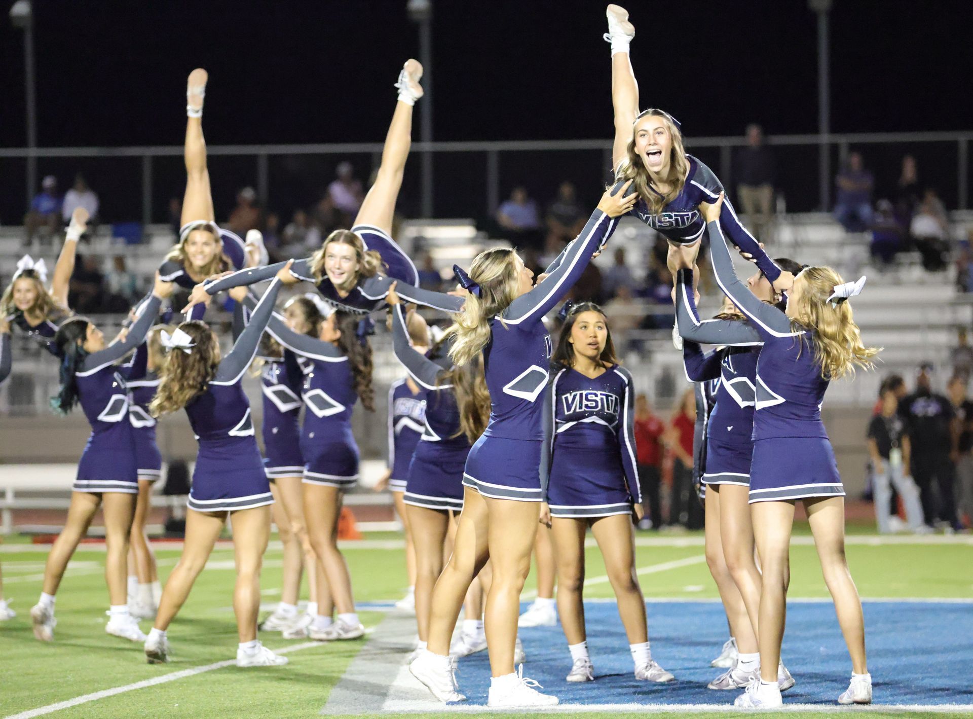Cheerleaders in blue uniforms performing a stunt on a football field at night Vista Athletic Boosters