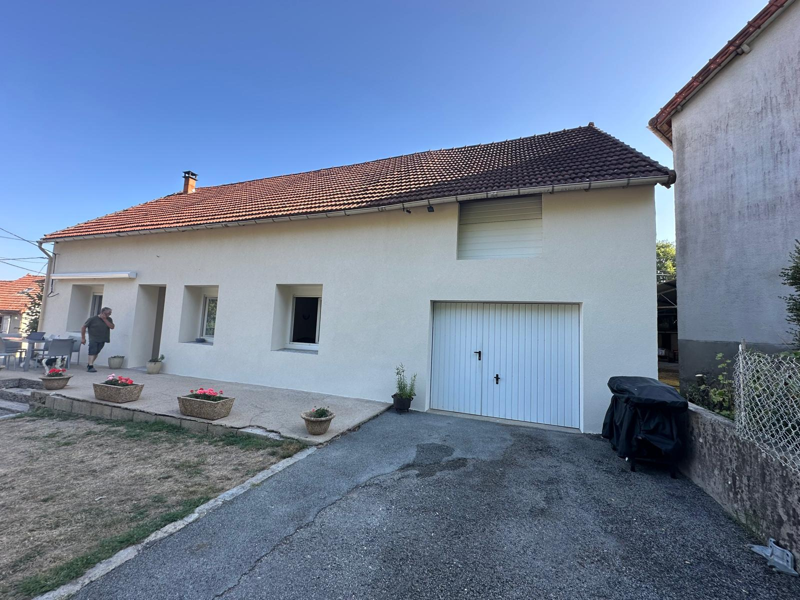 Maison beige avec allée et porte de garage. Une personne se tient près de la porte d'entrée, ciel bleu au-dessus.