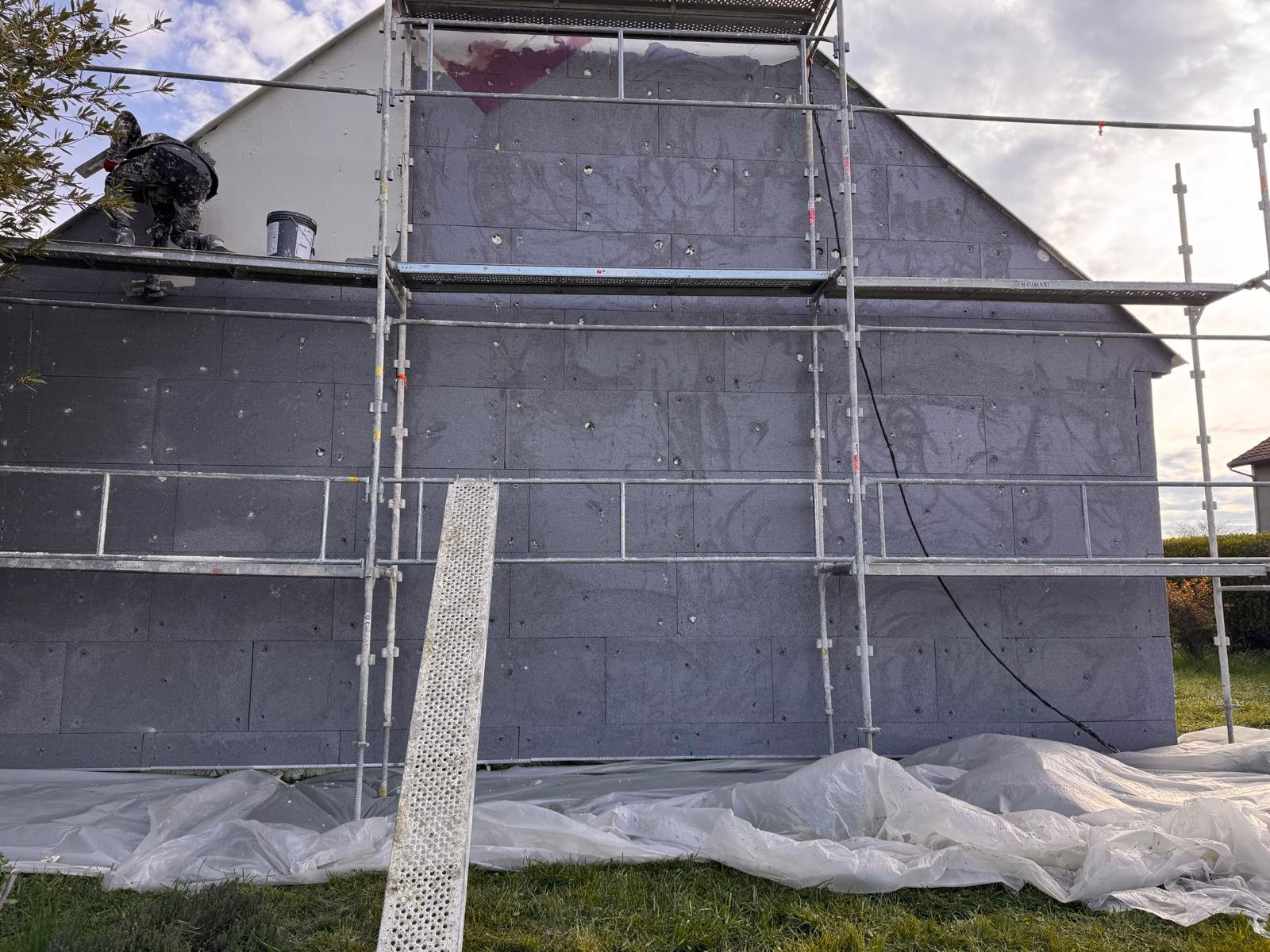Façade de maison en rénovation avec échafaudages ; ouvrier sur le toit, isolation grise, ciel couvert.