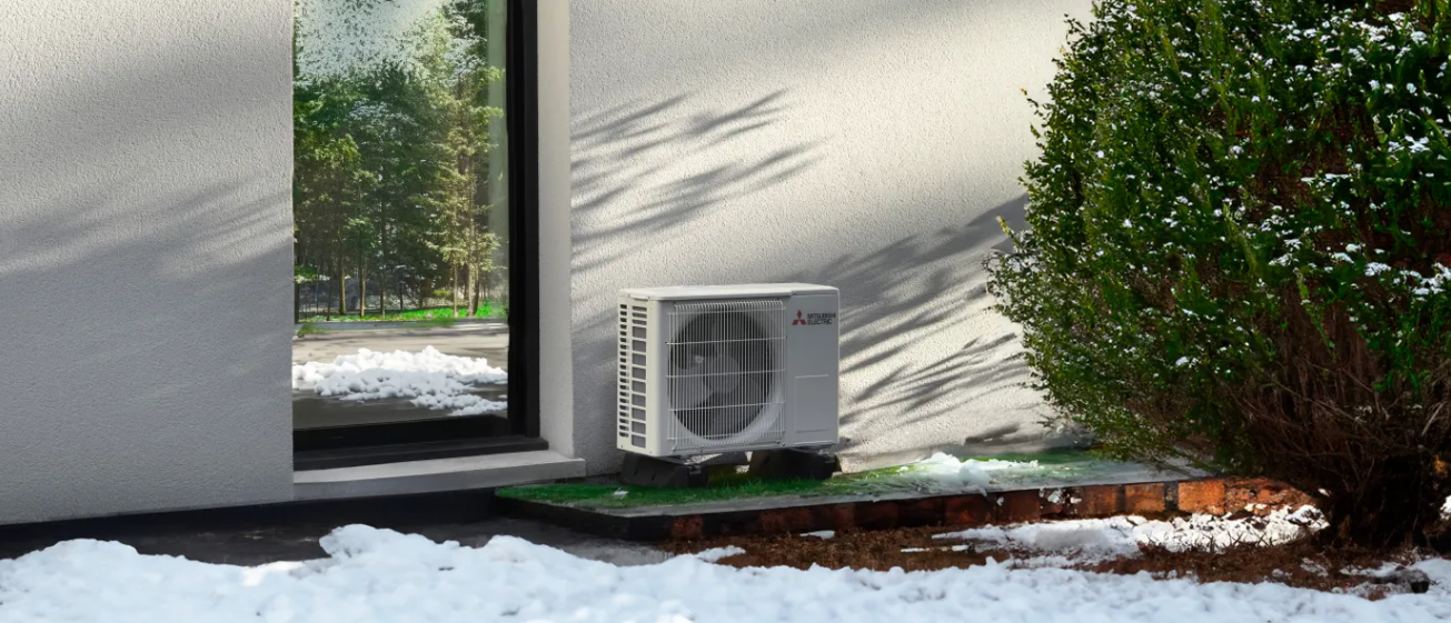 Un climatiseur extérieur est installé près d'une fenêtre enneigée. Un buisson vert est visible sur la droite.