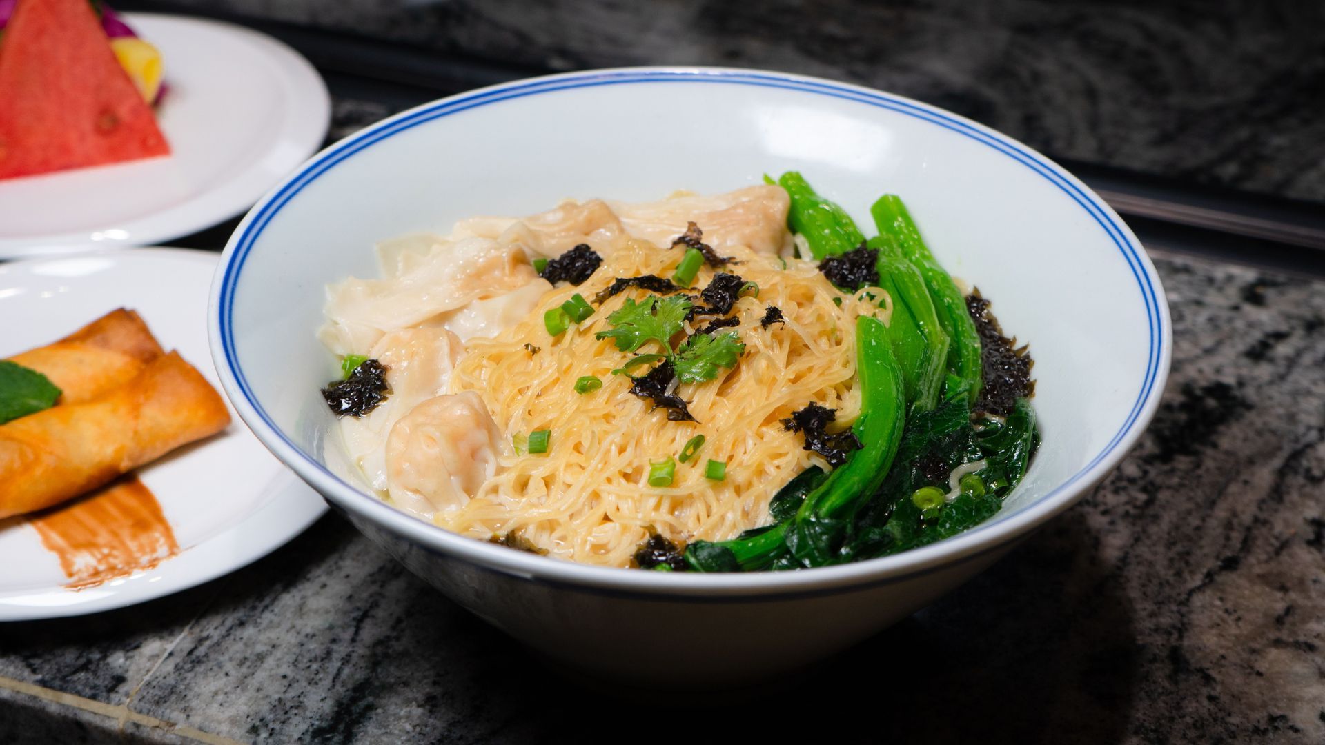 Bowl of creamy pasta with broccoli and black olives on a table, with spring rolls on a side plate.
