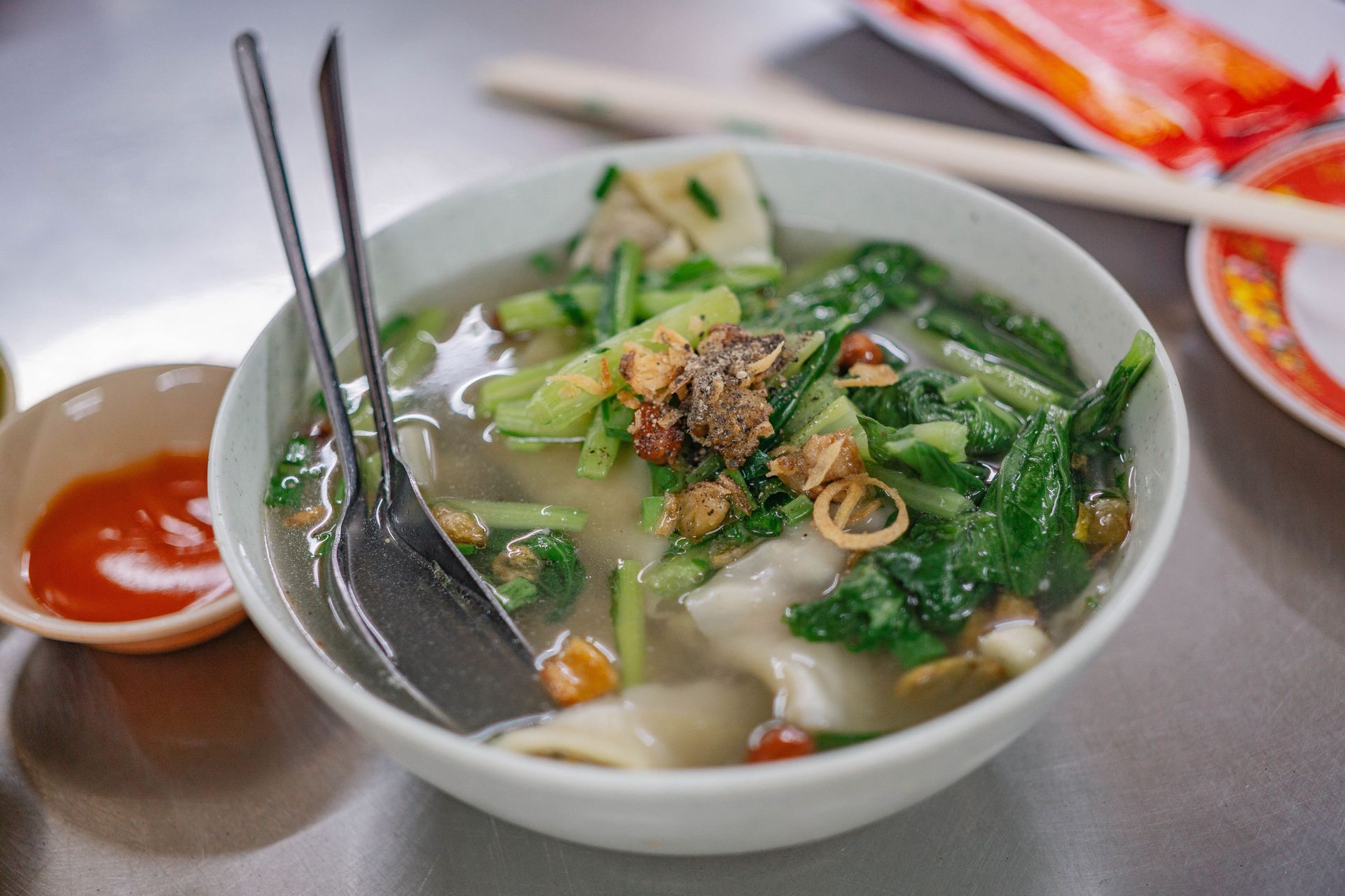 Bowl of noodle soup with greens and meat, served with chopsticks and a spoon on a table