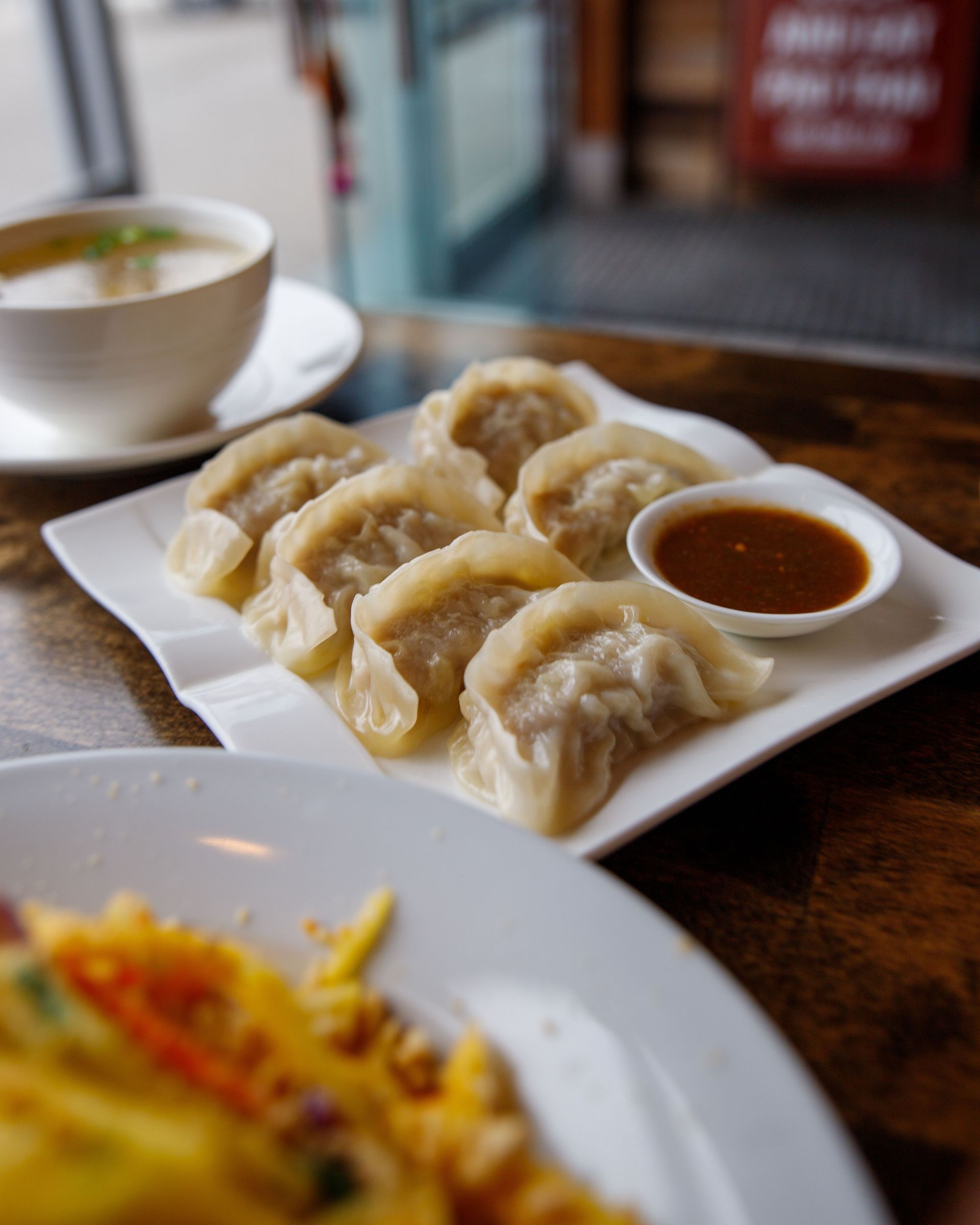Plate of steamed dumplings with dipping sauce on a wooden table, with soup and a blurred dish in the foreground