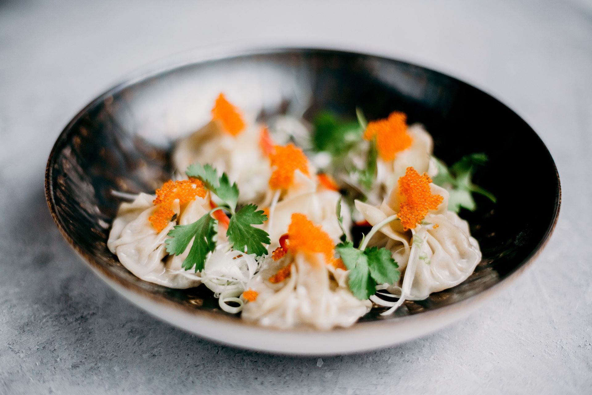 Black bowl of dumplings topped with cilantro, carrots, and sauce on a gray surface