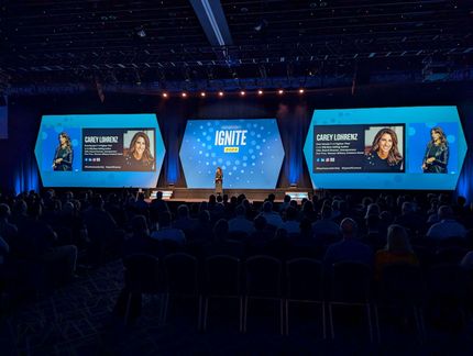 Large conference stage with blue screens, a central speaker, and an audience seated in the dark.
