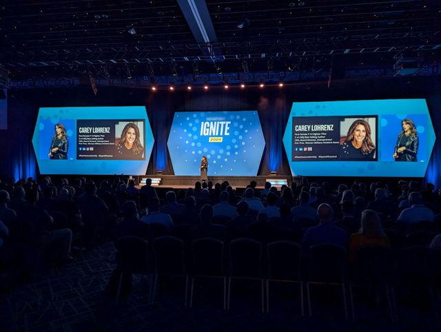 Large conference stage with blue screens, a central speaker, and an audience seated in the dark.