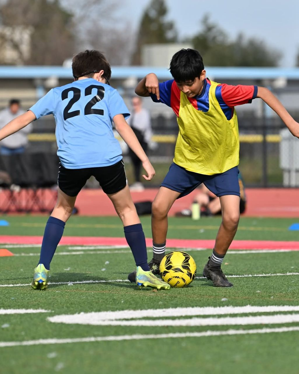 Youth soccer players in blue pinnies stand on a turf field while a coach in a dark blue tracksuit observes them.