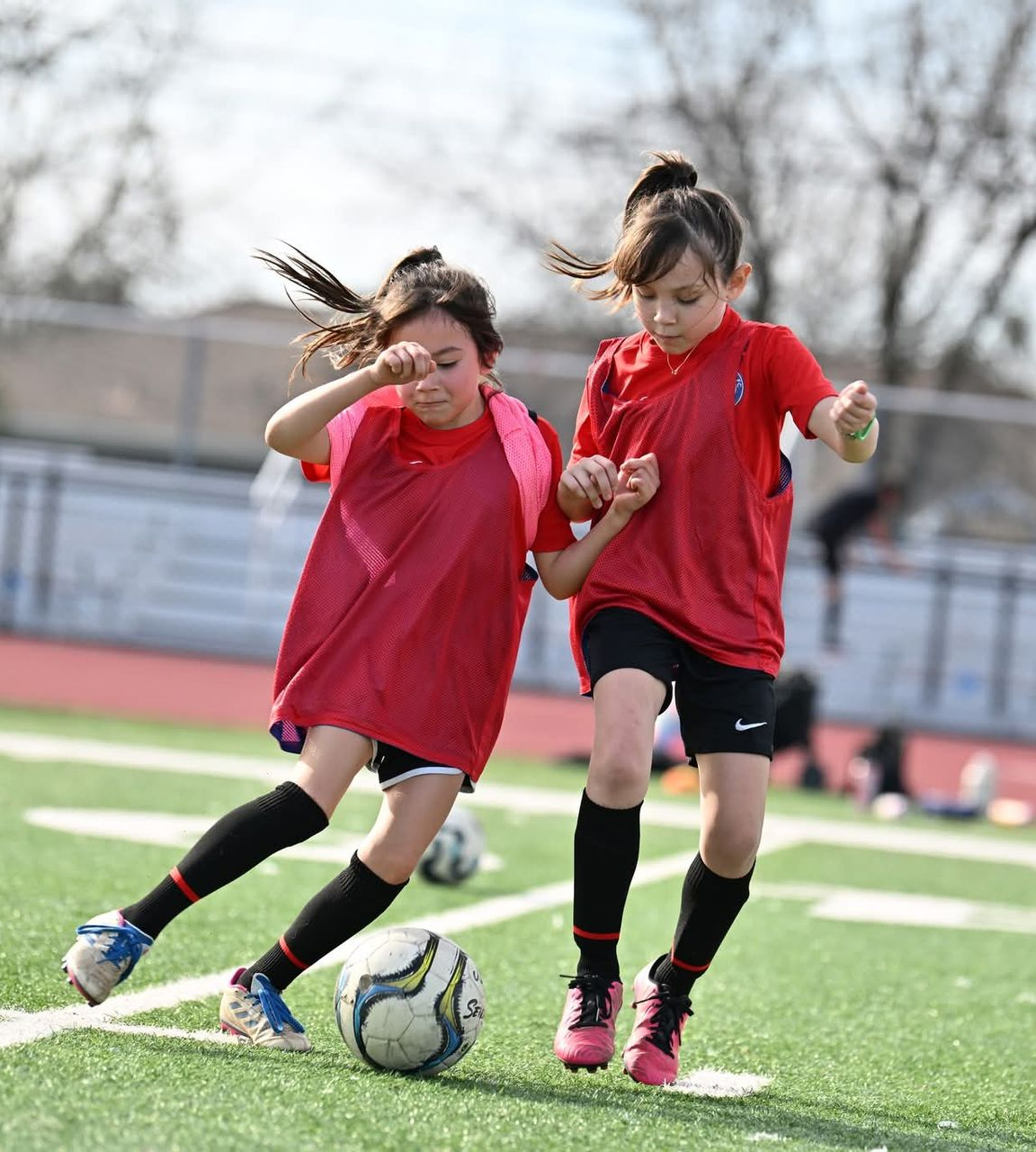 Two youth soccer players in red practice jerseys run together across a field while dribbling a soccer ball.