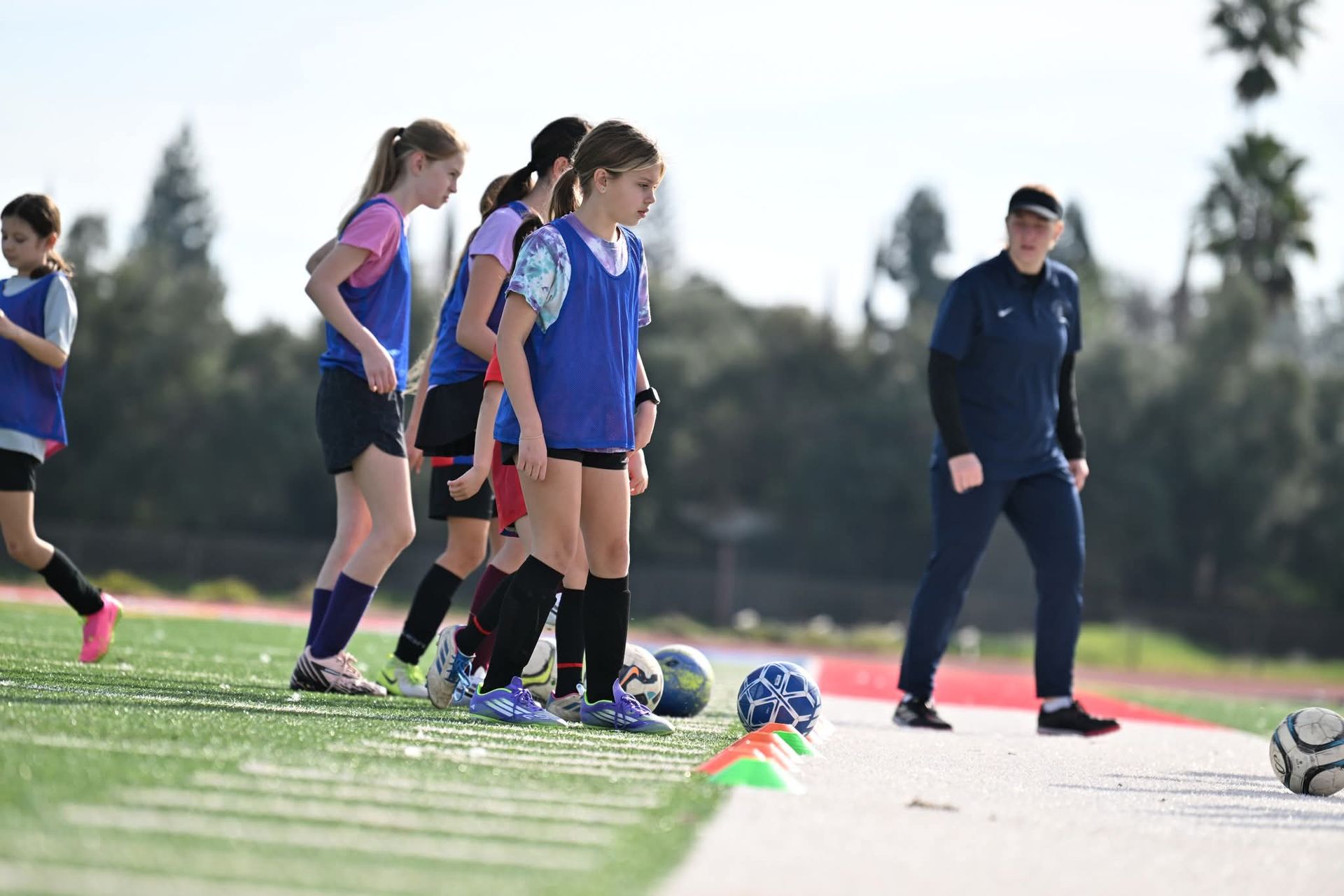 Youth soccer players in blue pinnies stand on a turf field while a coach in a dark blue tracksuit observes them.