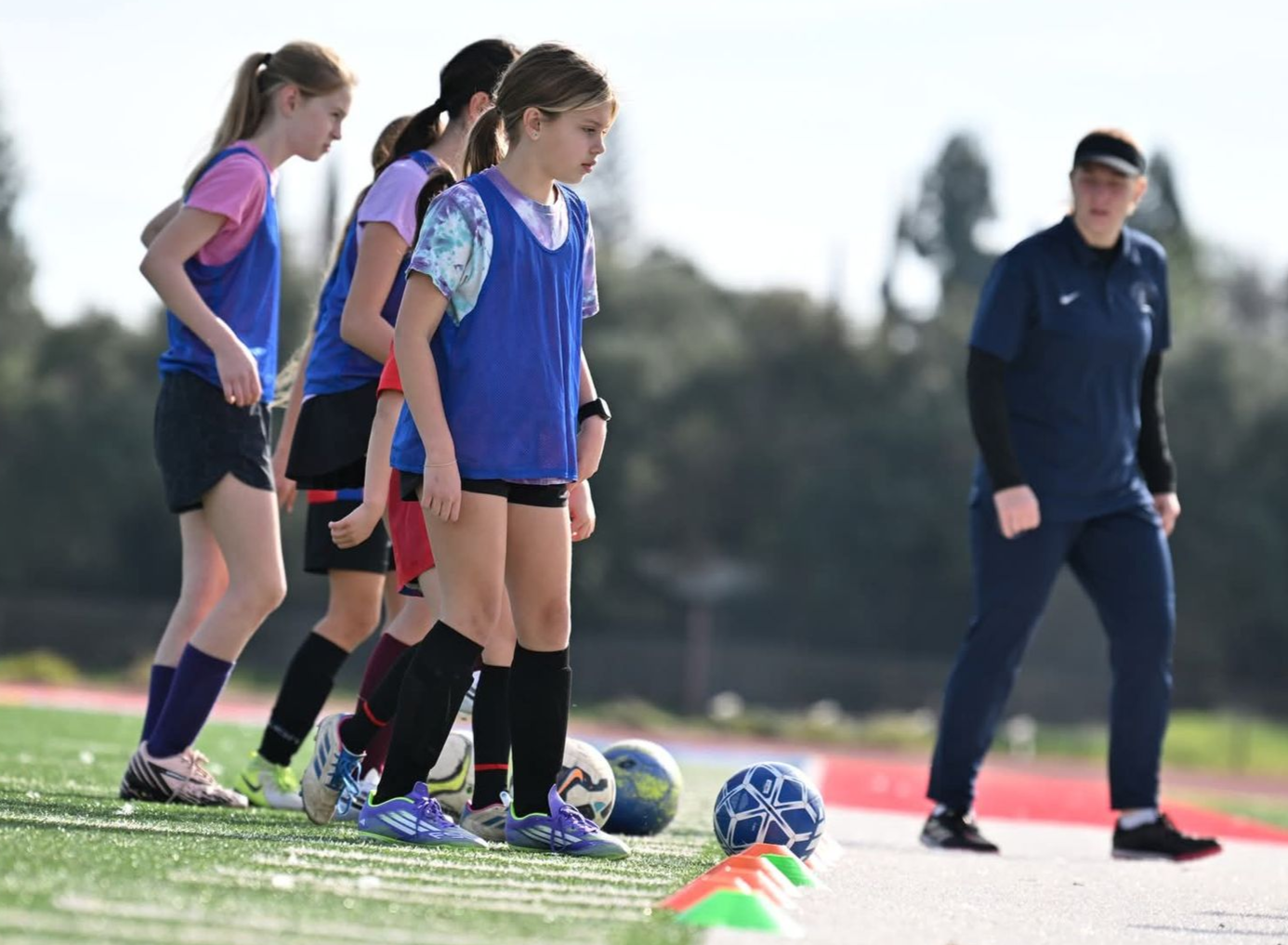 A coach stands near a line of players in blue training vests, who are positioned by soccer balls on an outdoor field.