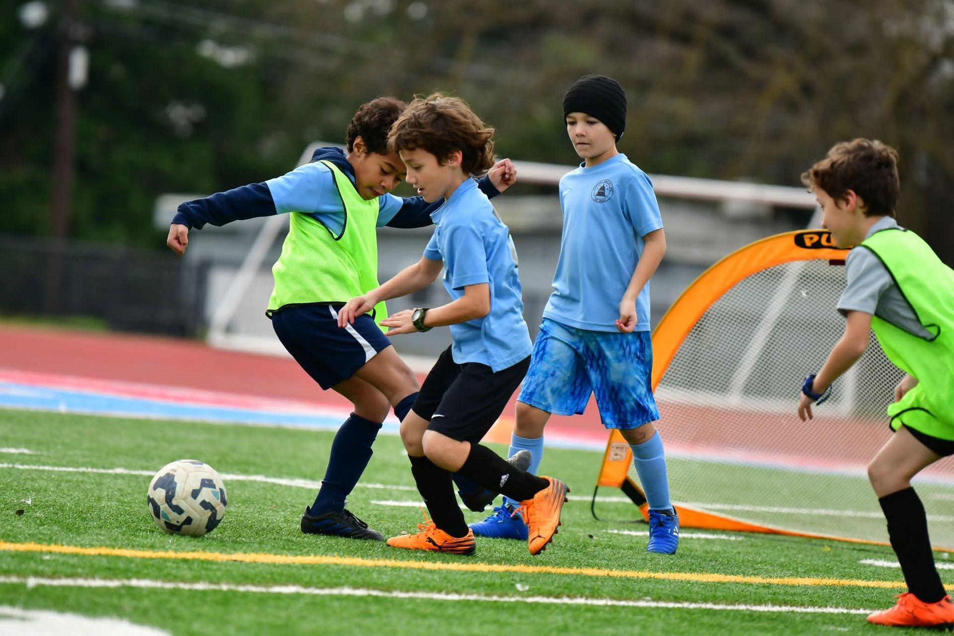 Four children in sports uniforms play soccer on an artificial turf field near a small portable goal.