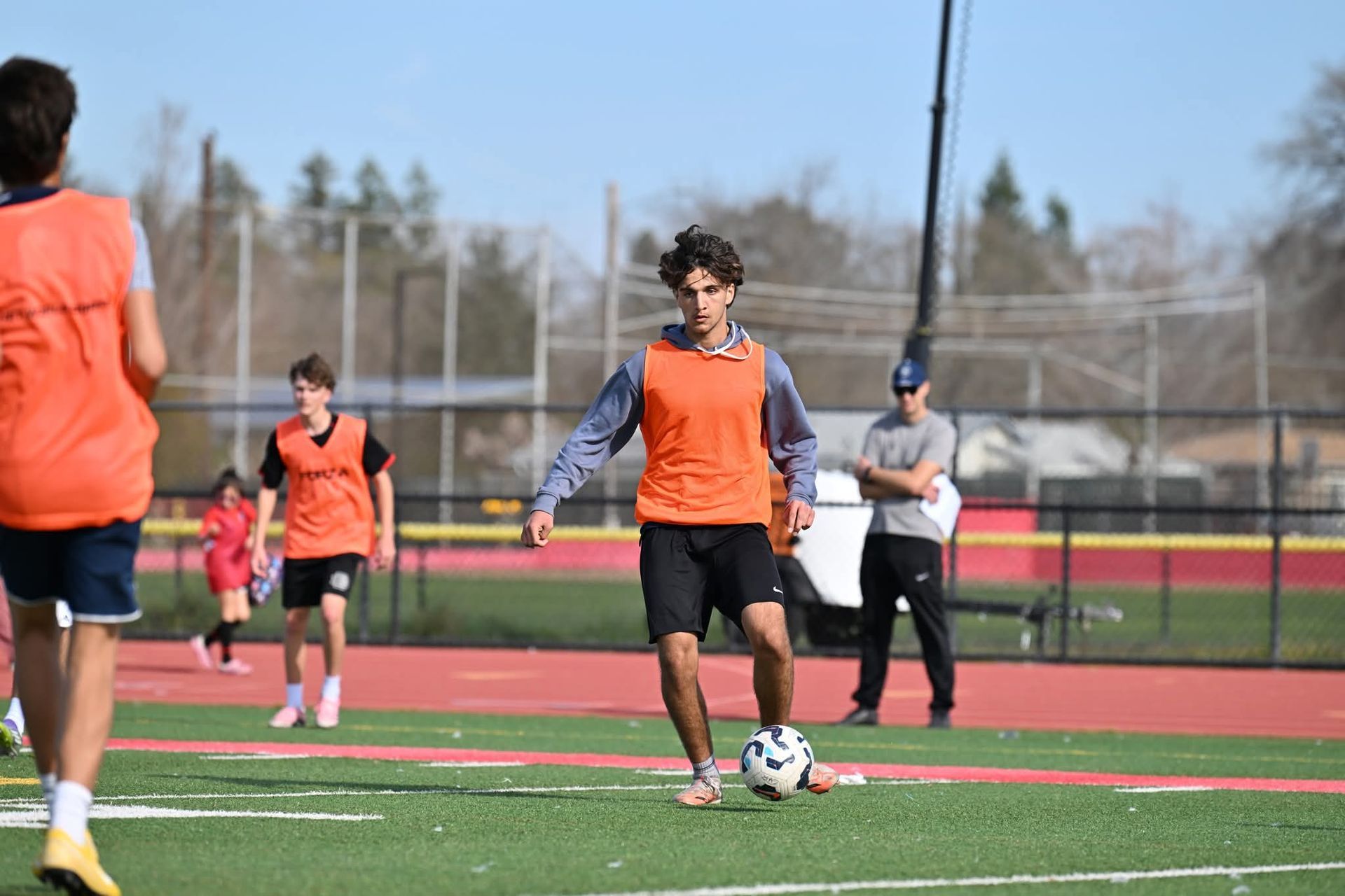 A person in an orange training bib and black shorts dribbles a soccer ball on a track field with others in the background.