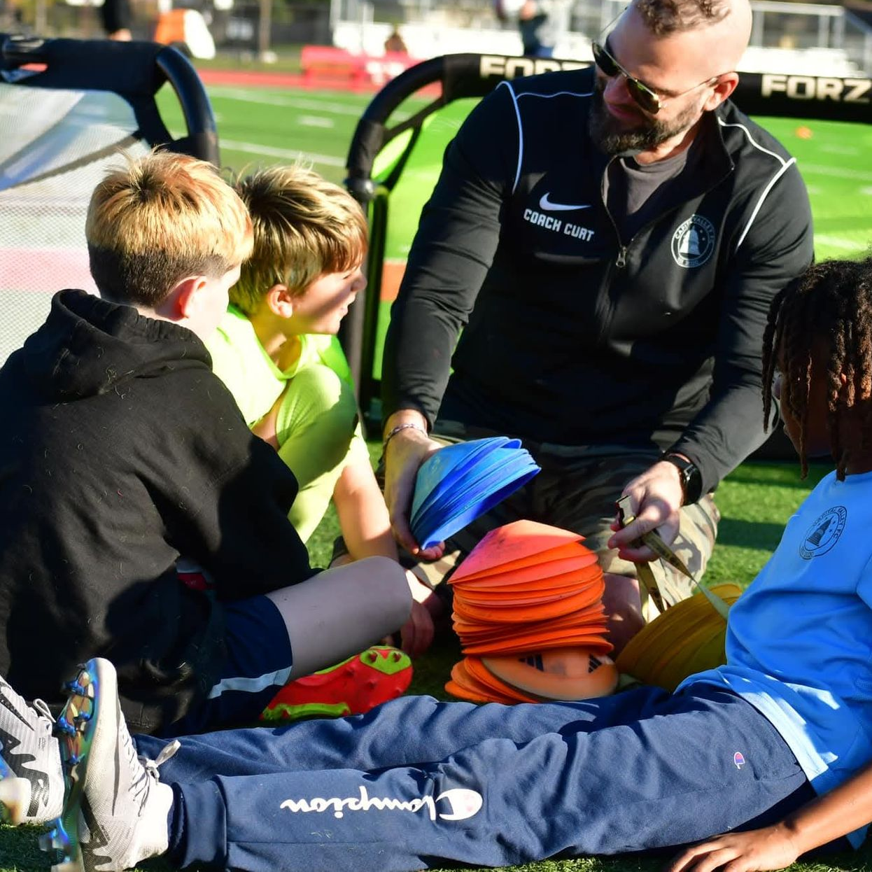 A coach sits with youth players on a field, holding a stack of orange training cones and explaining an exercise.