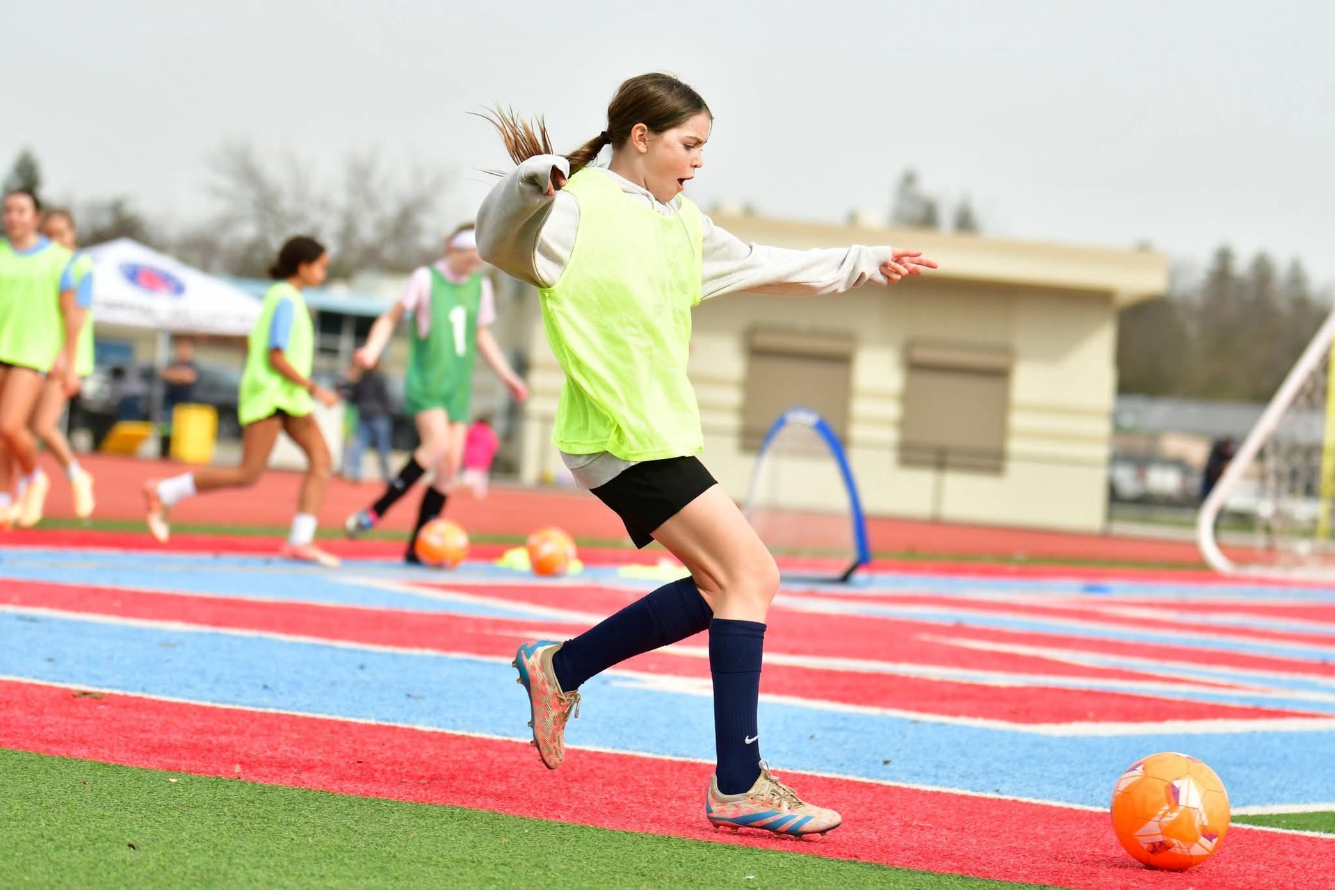 A soccer player in a yellow vest runs across a red and blue field, preparing to kick an orange ball during a practice.