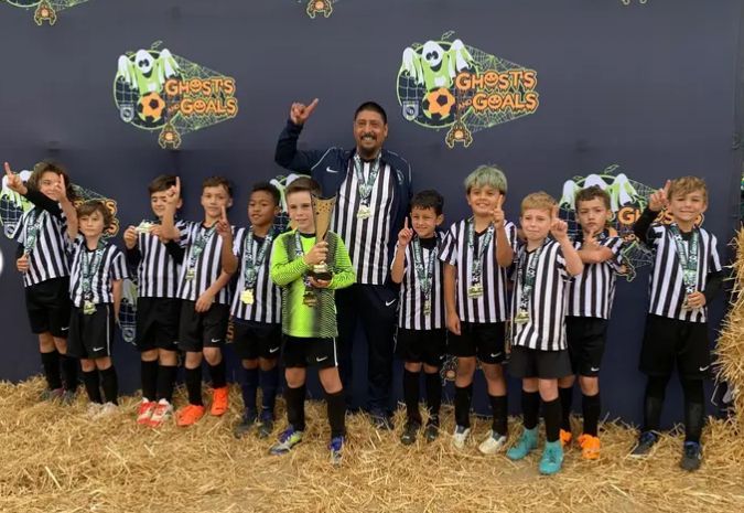 A youth soccer team and their coach stand smiling, wearing medals in front of a "Ghosts & Goals" tournament backdrop.