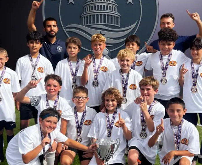 A group of youth athletes in white shirts and medals celebrating with a trophy in front of a sports logo banner.