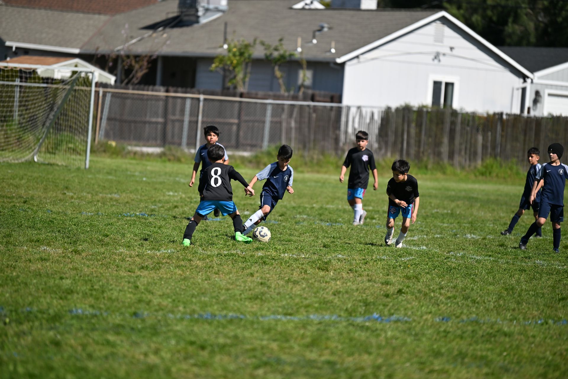 Young soccer players in matching jerseys compete for the ball on a grassy field in front of suburban houses.