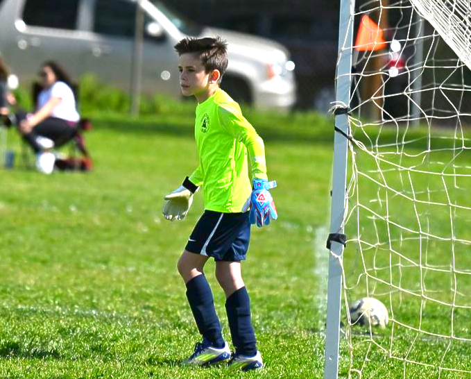 A goalkeeper in a bright neon yellow jersey and dark shorts stands on a grass field near a goal net.