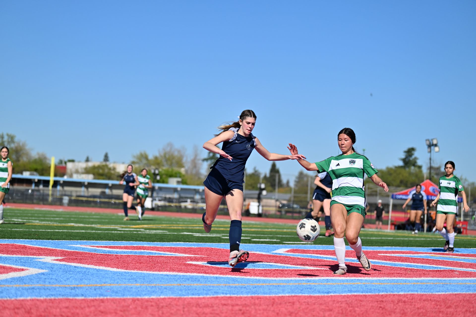 Two soccer players contest a ball on a blue and red field during an outdoor match on a sunny day.