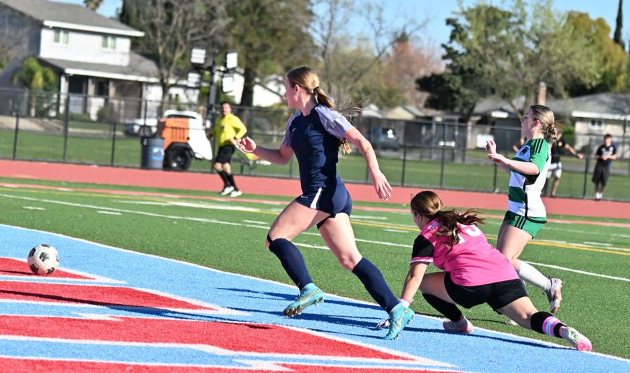 Three soccer players compete on a green field with a blue and red track, one pursuing a ball as another slides to defend.
