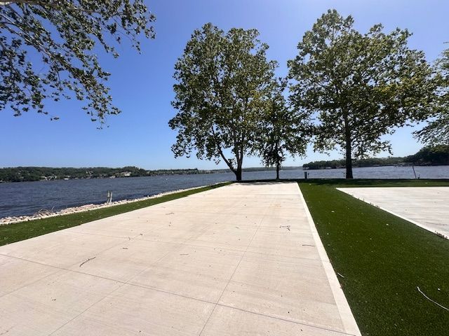 Concrete patio with artificial grass border by a lake, trees in background, sunny day.