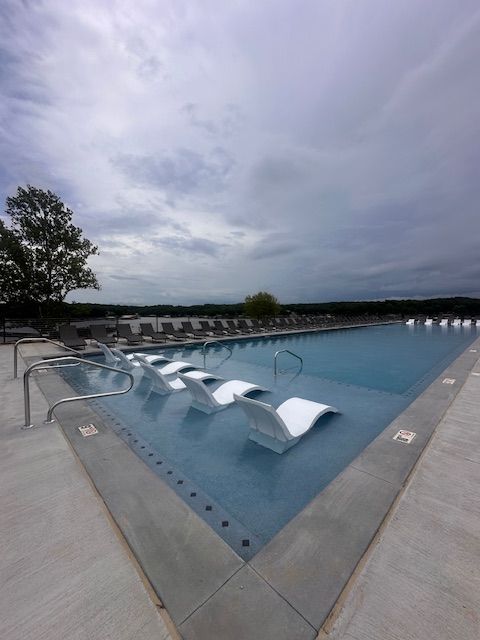 Outdoor pool with white in-water lounge chairs, cloudy sky.