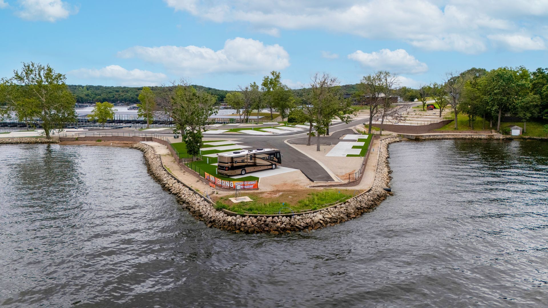 RV parked on a waterfront peninsula with stone breakwater. Trees and a building in the background.