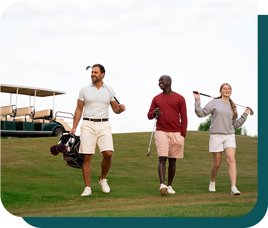 Three people are walking on a golf course with a golf cart in the background.