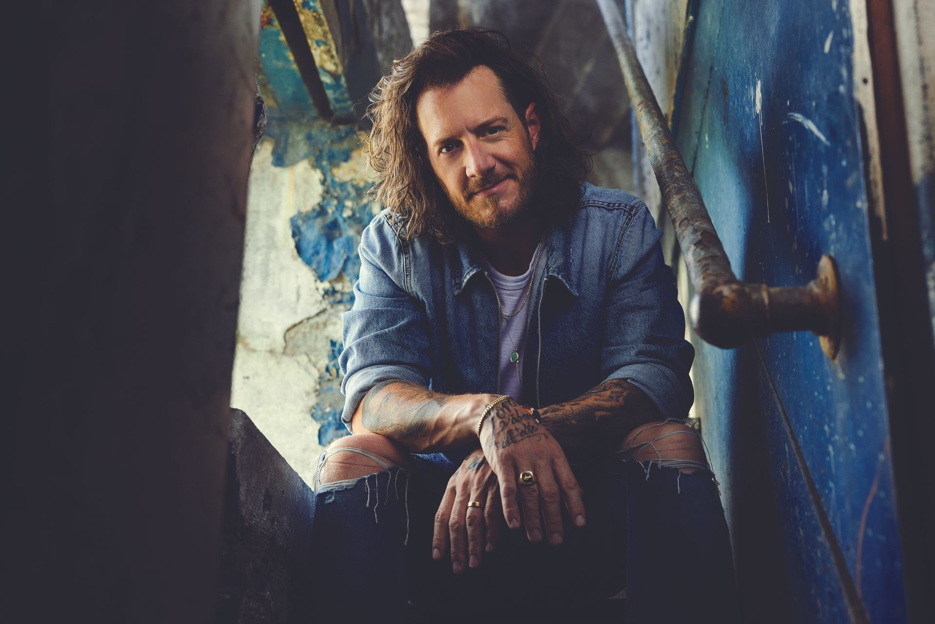 A man with curly hair and a beard is sitting on the ground in front of a blue wall.