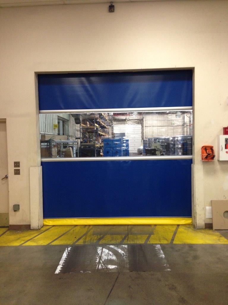 Blue industrial roll-up door with a window panel, set in a factory-like space. Yellow floor markings and some equipment are visible through the window.