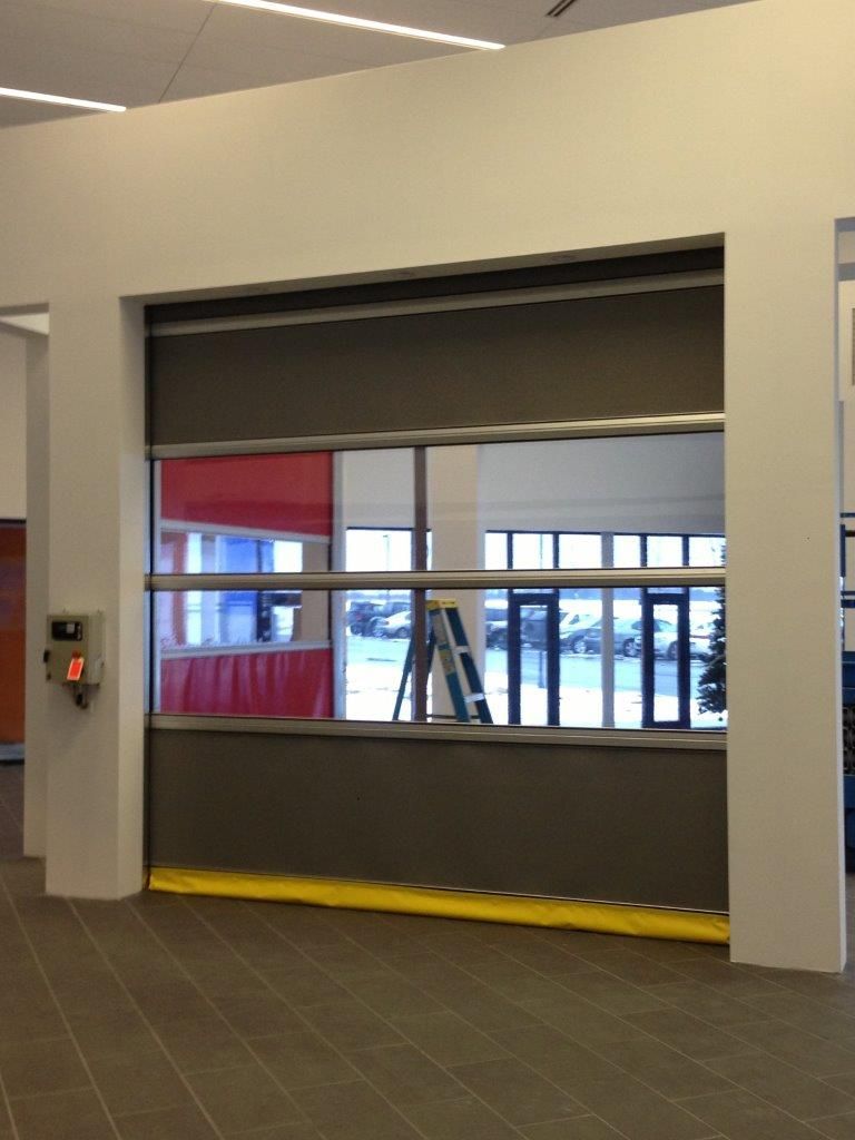 A high-speed industrial door with a gray roll-up door and a window, yellow safety strip, and a control panel on the left.