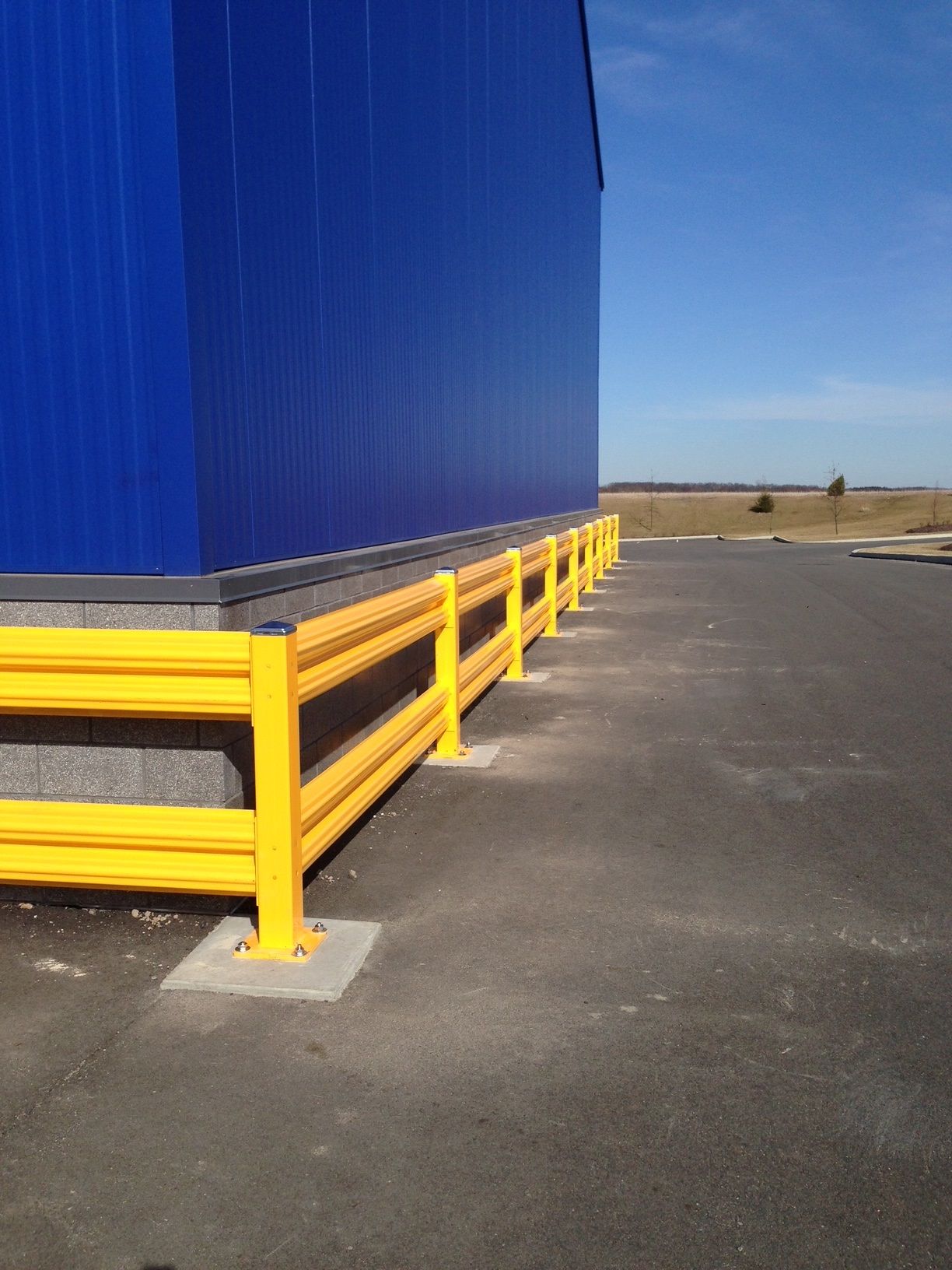 Yellow safety barriers line the side of a bright blue building, protecting an adjacent dark asphalt area under a clear sky.